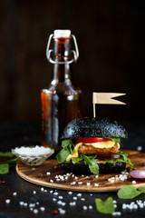 Black burger with cutlet, greens, cheese, onions and tomatoes and a bottle of beer on a wooden plate on a dark table