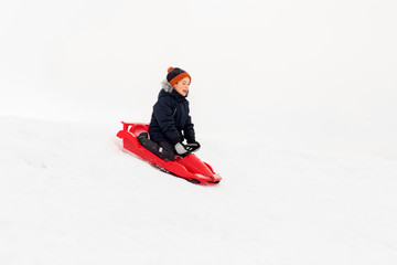 Fototapeta na wymiar childhood, sledging and season concept - happy little boy sliding on sled down snow hill outdoors in winter