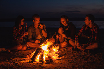 Camp on the beach. Group of young friends having picnic with bonfire. They eat sausages from fire