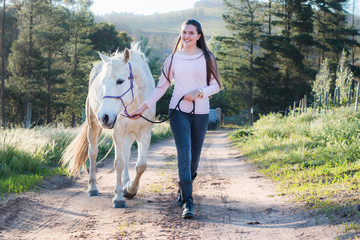 Teenage girl walking next to a white Boerperd horse on a dirt road, leading him with his head...