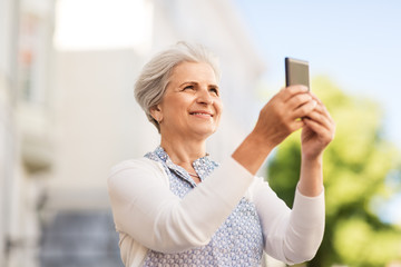 travel, tourism and technology concept - happy senior woman photographing by smartphone in city