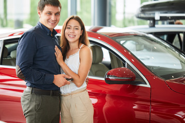 Buying their first car together. Young caucasian positive couple of successful businessman and his wife hugging near red glistering car in car dealership, man holds the key from their new car