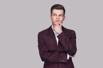 Portrait of serious thinking handsome young man in violet suit and white shirt, standing, touching his chin and looking away with thoughtful face. indoor studio shot, isolated on grey background.