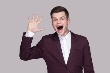 Hi, nice to see you. Portrait of funny handsome young man in suit and white shirt, standing, looking at camera with amazed face and greeting gesture. indoor studio shot, isolated on grey background.