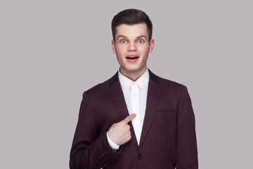 Am I winner?! Portrait of handsome amazed young man in violet suit, white shirt, standing, looking at camera, pointing himself with surprised face and asking. studio shot, isolated on grey background.