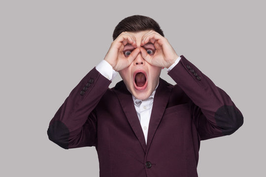 Portrait of amazed handsome young man in violet suit and white shirt, standing, looking at camera, shocked and screaming with binoculars gesture. indoor studio shot, isolated on grey background.