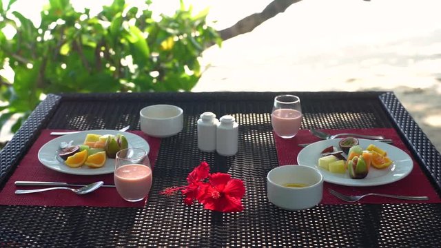 Early morning breakfast table on beach at seaside restaurant, plate with fresh fruit, juice and yoghurt bowls, hibiscus flowers. Slow motion 50p.