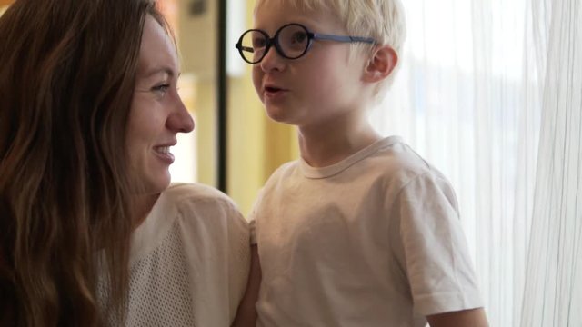 Beautiful young woman and her son are chatting. The boy tells a funny story, mom smiles. The child is like a little professor. The family is wearing white sweaters. They are sitting in a cafe and wait