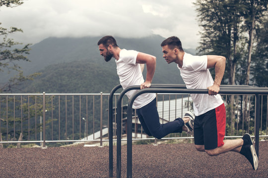 Two concentrated young sportsmen in white t-shirts doing dips on horizontal parallel bars outdoors, over sky and nature background