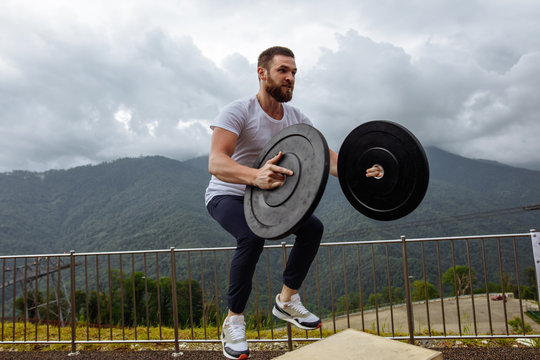 Handsome bearded athletic man in white t-shirt doing jumping squats on box with weights, plyometric exercise on top of the mountain. Outdoor functional training with cross-fit equipment.