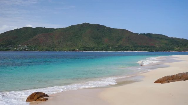 Early morning view from Curieuse beach onto Praslin, Seychelles (granitic island), waves crashing on white sand, sunny day. Medium shot. Slow motion 50p