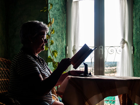 Portrait Of Senior Woman Reading A Book In A Rural House With Natural Sun Light