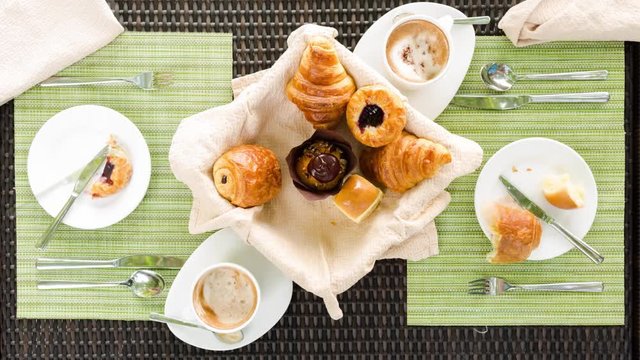 Overhead stop motion view of couple having coffee(foam cappuccino) and freshly baked pastries(croissants) for breakfast at table. 4K, 25p.