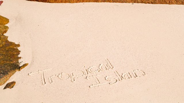 Tropical island stop motion writing text in sand on beach(tropical island) with towels, sun hat, sunglasses, bag and coconut. Seychells, Praslin, Anse Lazio. 4K, 25p.