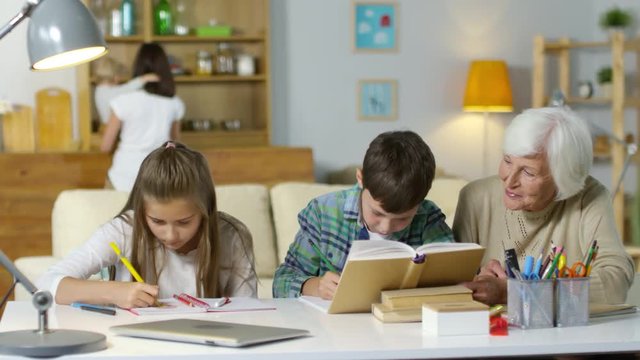 Brother and sister doing homework with help of grandmother at desk in the living room while mother walking with baby in the background