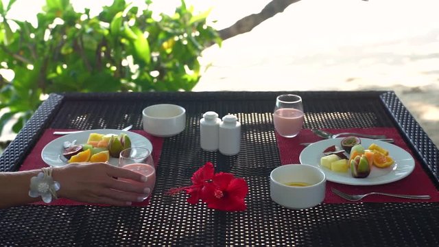 Close-up breakfast table on beach seaside restaurant, couple(hands) drinking juice, plate fresh fruit, yoghurt bowls, hibiscus flowers. Slow motion 50p.