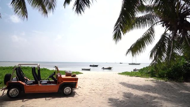 Slow linear dolly shot of tourist beach buggy/Moke parked on tropical island beach, white sand, palm trees and boats on sea. Medium shot, Slow motion 50p.