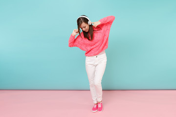 Full length portrait of happy young woman in knitted rose sweater, white pants, headphones isolated on bright pink blue pastel wall background in studio. Fashion lifestyle concept. Mock up copy space.