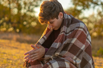 young frozen teenager covered by a warm blanket sitting on the grass field against the sun а