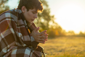 young frozen teenager covered by a warm blanket sitting on the grass field against the sun а