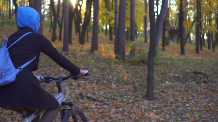 Muslim woman in a hijab and with a backpack on her back rides a bicycle in autumn park