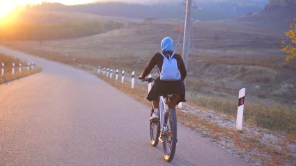 Muslim woman in hijab with backpack riding a bike fast on the road beautiful sunset, rear view