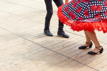 Legs of male and female rock 'n roll dancers on a wooden floor with copy space. The man in black pants and black shoes, the woman in a wide red skirt with roses and white dots on black.