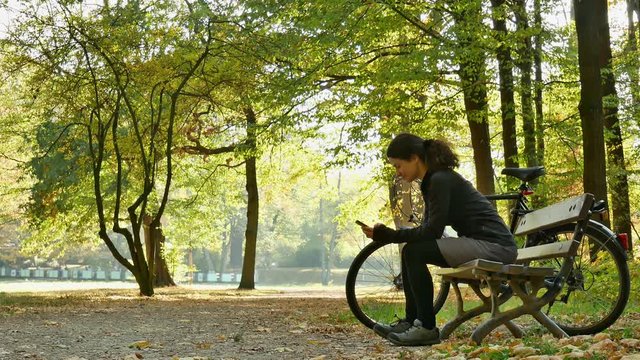 City park: Female cyclist resting on a park bench observing something than reading on her phone.(Germany)