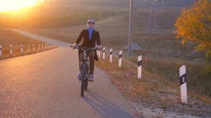 Muslim woman in hijab with backpack riding a bike on the road setting sun,slow mo