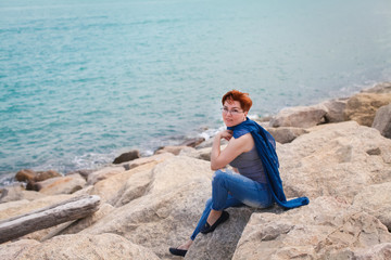 Fototapeta na wymiar Adult caucasian women sitting on rocky beach with blue neckchief relaxing and thinking about something
