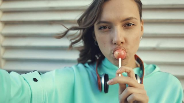 Pretty smiling girl licking lollipop and making selfie using smartfone. Close-up portrait young beautiful caucasian woman holding phone taking self photo. Slow motion video