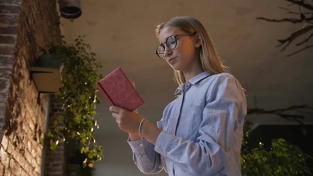 Beautiful girl in glasses making some notes in the notebook at indoors sitting on a table opposite the brick wall in the evening. Beautiful young woman sits on the table with a notepad and pen in