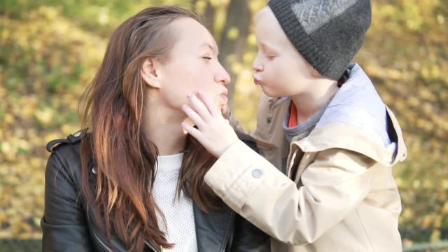 the boy and his mother are walking in the autumn park. They sit on fallen leaves. Son kisses his mother on the lips. The woman pulls away and laughs. The boy is trying to kiss her again. Cute family