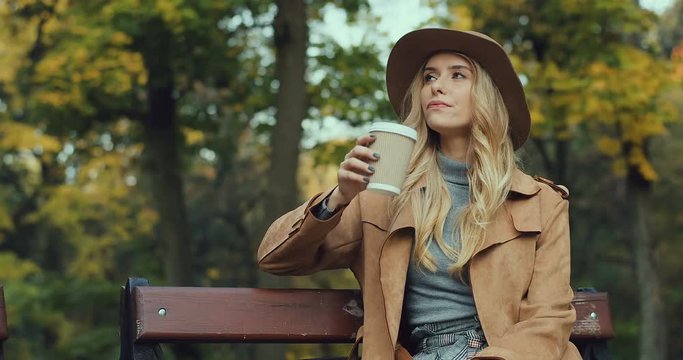 Caucasian blonde attractive and stylish young Caucasian woman sitting on the bench in a park, drinking coffee while waitingfor somebody and looking at the watch on her hand.