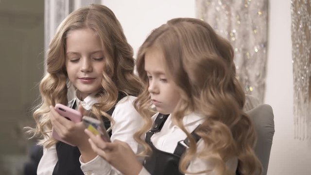 A side view of two charming fair-haired girls holding phones and chatting. One girl mimes V sign and says something; the second one points to the screen, and keeps on talking.