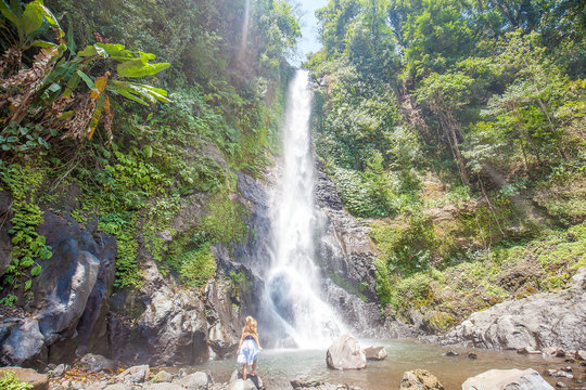 Young woman with long hair standing back on the stones in front of Gitgit waterfall among green tropical trees on the Bali island, Indonesia