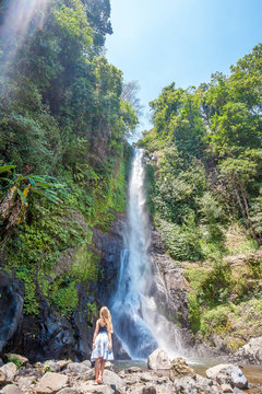 Young woman with long hair standing back on the stones in front of Gitgit waterfall among green tropical trees on the Bali island, Indonesia