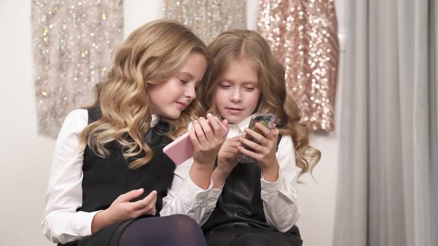 Two pretty fair-haired girls are sitting on the couch indoors. They both hold phones and look at one screen. The girl to the right says something and the girl to the left scrolls her screen.