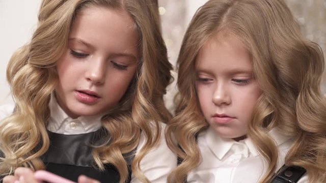 A close up of two fair-haired little girls. They are looking down at phones' screens and talking.