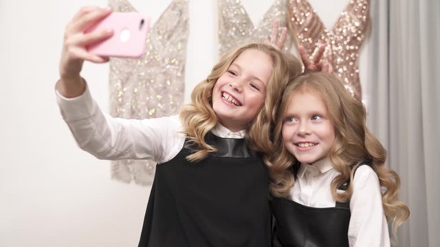 Two lovely girls with long curly fair hair are taking a selfie. The older one holds a phone in front of them. They make V signs, pull funny faces, show tongues and make bunny ears.