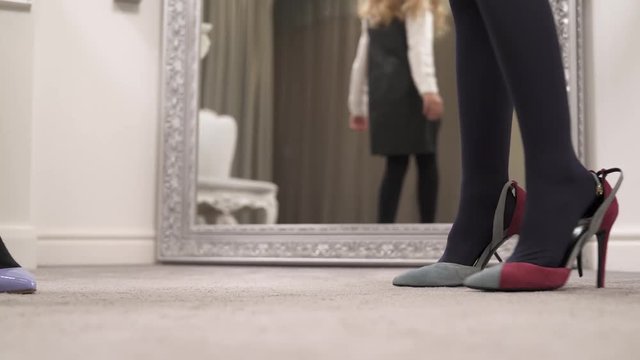Girl's legs in black tights wearing high-heeled shoes and walking around the room in front of the mirror. Another girl comes and tries walking from the right to the left in shot.