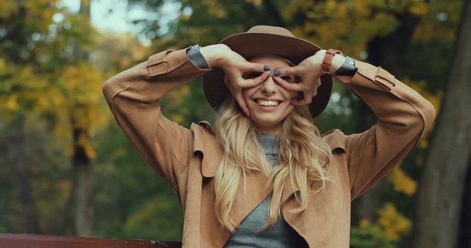 Portrait shot of the pretty cute blond young woman in a hat and coat laughing while posing to the camera and doing funny muzzles and gestures. Fall in the park.