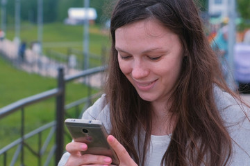 Young brunette woman chatting, typing a message on mobile phone walking along the promenade in the city.