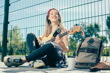 Young lady feeling happy while sitting near the chain link fence and playing the guitar
