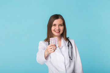 Attractive female doctor showing pink pills. Beautiful young physician holding medicaments over blue background.
