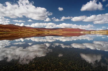 Obraz premium Colorful highland steppe desert shore of lake with dry yellow grass on the background of rocky mountains under clouds with mirror reflections