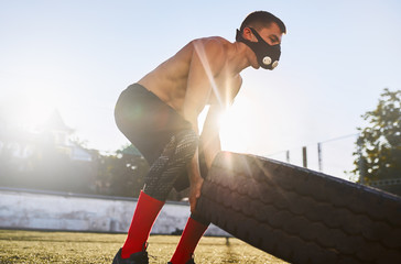 Side view shot of muscular male in mask to increase load on breathing muscles doing tire-flip exercises outdoors in stadium. Copy space for your advertising. Shirtless sportsman doing workout outside