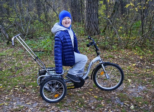 child autistic with disabilities on a tricycle with management for mom, in a blue cap and jacket in the Park for a walk
