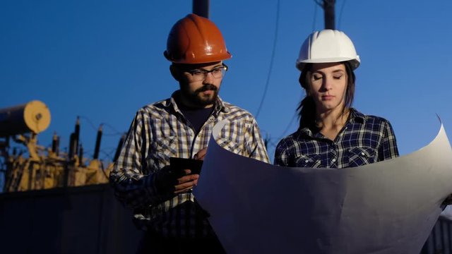 Woman in the male profession. Two engineers make calculations on a big piece of paper, looking around and checking information on tablet