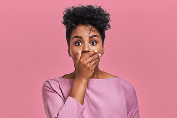 Horizontal shot of surprised African American woman covers mouth with both hands, tries to be...